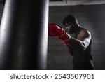 young strong african american boxer in red gloves hits punching bag, muscular athlete trains punch in dark gym and does sports on black background