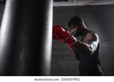 young strong african american boxer in red gloves hits punching bag, muscular athlete trains punch in dark gym and does sports on black background - Powered by Shutterstock