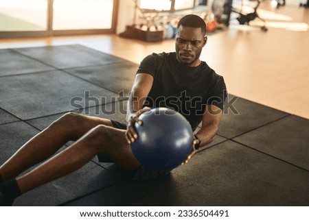 Young strong African American male athlete in black sportswear sitting on mat, doing Russian twist exercise with heavy medicine ball during fitness workout in gym