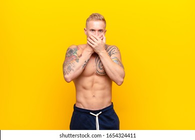 Young Strong Blonde Man Covering Mouth With Hands With A Shocked, Surprised Expression, Keeping A Secret Or Saying Oops Against Yellow Wall