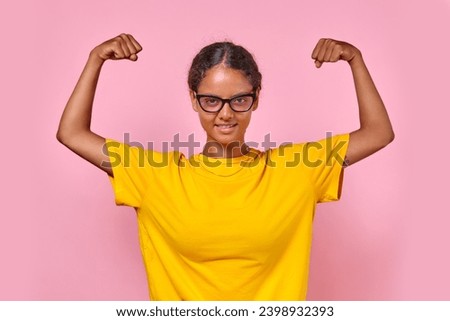 Young strong brave Indian woman teenager takes strongman pose and shows biceps to demonstrate independence and readiness for beginning of adulthood stands on plain pink background.
