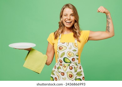 Young strong cool happy housewife housekeeper chef cook baker woman wears apron yellow t-shirt hold in hand empty plate towel show hand biceps isolated on plain green background. Cooking food concept - Powered by Shutterstock