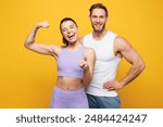 Young strong fitness trainer sporty two man woman wear blue clothes spend time in home gym show hand biceps muscles point camera on you isolated on plain yellow background. Workout sport fit concept