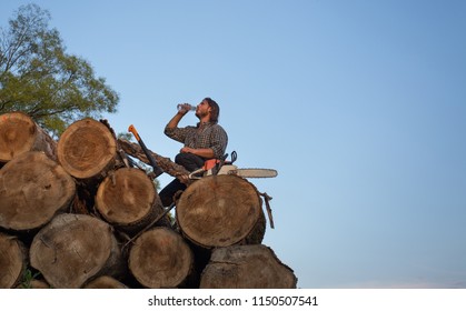 Young strong lumberjack sitting on pile of trunks with chainsaw and ax beside and drinking water from bottle - Powered by Shutterstock