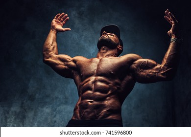 Young strong man bodybuilder in cap on wall background. Dark dramatic colors.