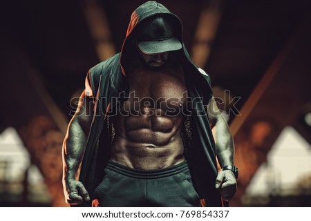 Young strong man bodybuilder with informal style clothing with hood standing in urban city interior. Dark red colors.