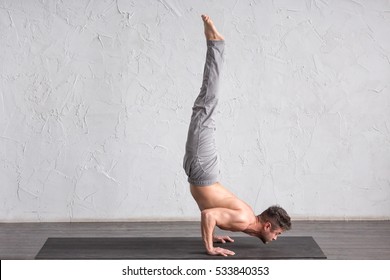 A young strong man doing yoga exercises - handstand. Studio shot over white brick background and black floor