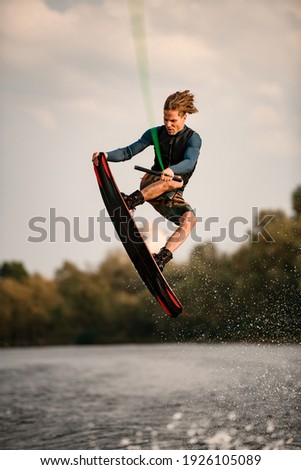 young strong man masterfully jumping over river water on wakeboard. Sky and green trees in background