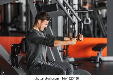 Young Strong Muscular Fit Man Back Muscles Workout Training With Weights On The Machine In The Gym. The Athlete Listens To Music In Earphones.