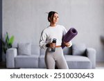 Young strong sporty athletic fitness trainer instructor woman wearing green tracksuit holding in hand yoga mat drink water training do exercises at home gym indoor. Workout sport motivation concept