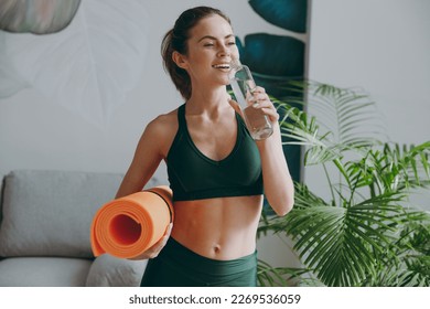 Young strong sporty athletic fitness trainer instructor woman wearing green tracksuit holding in hand yoga mat drink water training do exercises at home gym indoor. Workout sport motivation concept
