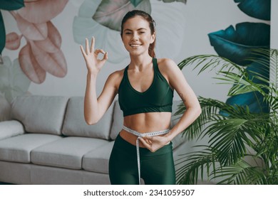 Young strong sporty athletic fitness trainer instructor woman wears green tracksuit hold measure tape on waist show ok okay training do exercises at home gym indoor. Workout sport motivation concept - Powered by Shutterstock