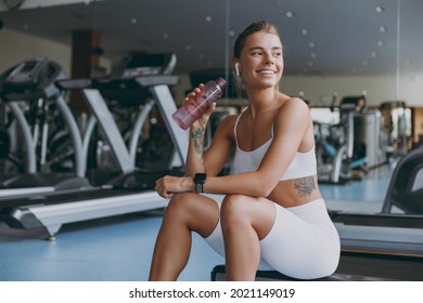 Young strong sporty athletic sportswoman woman in white sportswear earphones listen music warm up training sitting near treadmill drink water look aside in gym indoor Workout sport motivation concept. - Powered by Shutterstock