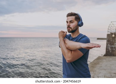 Young strong sporty athletic toned fit sportsman man in sports clothes headphones warm up training do stretch hand exercise at sunrise sun over sea beach outdoor on pier seaside in summer day morning.