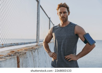 Young strong sporty athletic toned fit sportsman man 20s in sports clothes warm up training mobile cell phone stand akimbo at sunrise sun over sea beach outdoor on pier seaside in summer day morning. - Powered by Shutterstock