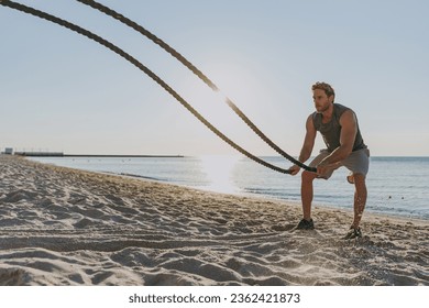 Young strong sporty athletic toned fit sportsman man in sports clothes warm up train work out with battle ropes at sunrise sun dawn over sea beach outdoor on sand beach seaside in summer day morning - Powered by Shutterstock
