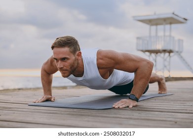 Young strong sporty athletic toned fit sportsman man wears sports clothes do push-ups exercises on floor warm up train at sunrise sun dawn over sea beach outdoor on pier seaside in summer day morning - Powered by Shutterstock