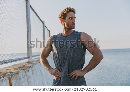 Young strong sporty athletic toned fit sportsman man in sports clothes warm up train stand akimbo arms on waist look aside at sunrise sun over sea beach outdoor on pier seaside in summer day morning.