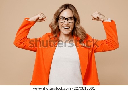Young strong sporty fitness employee business woman corporate lawyer 30s wear classic formal orange suit glasses work in office showing biceps muscles on hand isolated on plain beige background studio