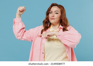 Young Strong Sporty Fitness Redhead Chubby Overweight Woman 30s Curly Hair In Casual Pink Shirt Point Finger On Biceps Muscles On Hand Demonstrate Strength Power Isolated On Pastel Blue Background.