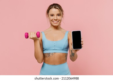 Young strong sporty fitness trainer instructor woman wear blue tracksuit spend time in home gym hold female dumbbells blank screen mobile phone isolated on plain pink background. Workout sport concept