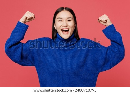 Young strong sporty woman of Asian ethnicity she wear blue sweater casual clothes show biceps muscles on hand demonstrating strength power isolated on plain pastel pink background. Lifestyle concept