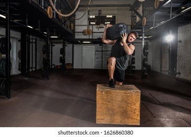 Young strong sweaty fit muscular man with big muscles holding heavy sandbag on his shoulder with his hands and climb on the jump box for legs cross workout training in the gym