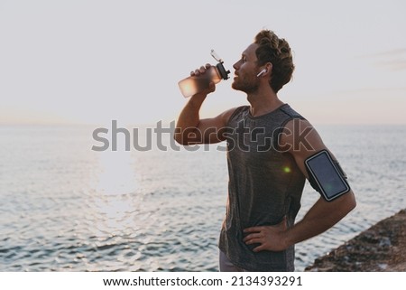 Young strong toned fit sportsman man in sport clothes earphones mobile cell phone warm up train drink stand akimbo water at sunrise sun over sea beach outdoor on pier seaside in summer day morning.
