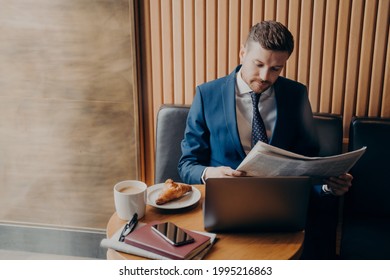 Young Stubbly Man Entrepreneur In Formal Wear Focused On Reading Financial Chronicle In Newspaper During Coffee Break While Enjoying His Cappuccino And Croissant, Sitting At Table With Open Laptop