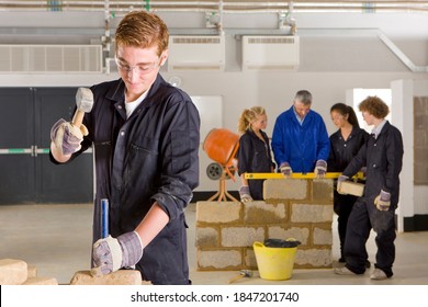 A young student in blue uniform hammering chisel on a brick in vocational school while his teacher training other students in the back ground