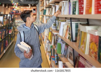 Young student boy standing in library holding books and reaching one for another
