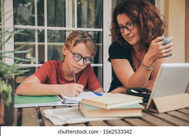 Young student doing homework at home with school books newspaper, digital pad helped by his mother. Mum control, help and teaching his son. Education, family, lifestyle and homeschooling concept.