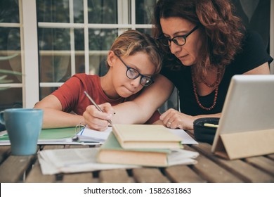 Young student doing homework at home with school books, newspaper and digital pad helped by his mother. Mum writing on the copybook teaching his son. Education, family lifestyle, homeschooling concept