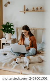Young student e-learning on laptop at home, cute small dog besides. Businesswoman working remotely, sitting on bed. Homeoffice for single woman living alone.