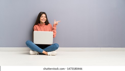 Young Student Girl With A Laptop On The Floor Pointing Finger To The Side