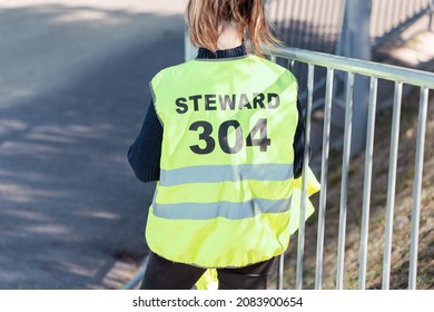 Young Student Girl Participates As A Volunteer In Ensuring The Security Of A City Event. Standing Near The Fence With The Inscription Steward On The Jacket