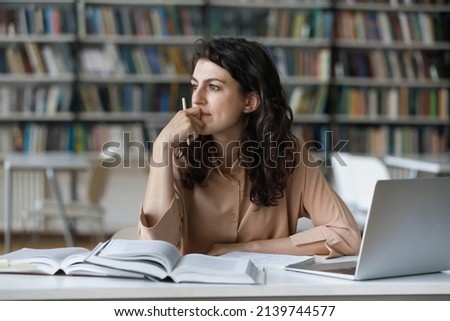 Young student girl sit at table with textbooks and laptop staring aside, studying alone in library, looks pensive and thoughtful search solution, prepare for exam, makes task feels confused or puzzled