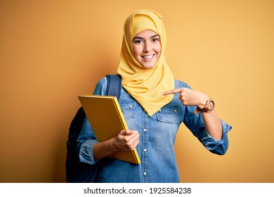 Young Student Girl Wearing Muslim Hijab And Backpack Holding Book Over Yellow Background With Surprise Face Pointing Finger To Himself