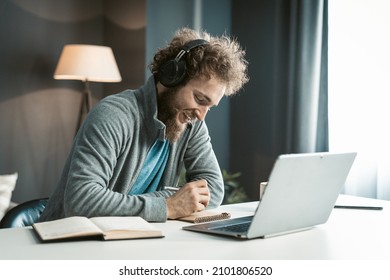 Young Student Has An Online Video Lecture Uses A Laptop And Headphones, Takes Notes In A Notebook. Curly-Haired Guy Sits In Earphones At The Table In His Office. Close-up. High Quality Photo