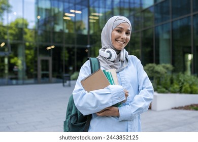 A young student in a hijab, headphones around neck, holding books, standing proudly in front of a campus building. - Powered by Shutterstock