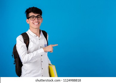 Young student holding yellow book carrying backpack points with on finger smiles on a blue background.