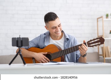 Young student learning to play guitar during self-isolating at home or online tutor. Smiling guy looks at guitar and makes video lesson for followers in living room during virus outbreak, free space - Powered by Shutterstock