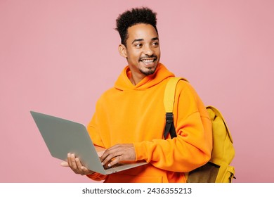 Young student IT man of African American ethnicity yellow hoody casual clothes backpack bag hold books use laptop pc computer look aside isolated on plain pastel pink background. High school concept - Powered by Shutterstock