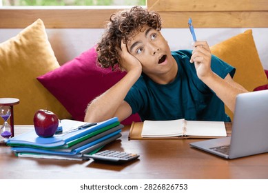 Young student procrastinating while studying at home, looking distracted and overwhelmed by homework, with laptop, notebooks, calculator, apple and hourglass on the desk. Concept of education, stress, time management and lack of focus.