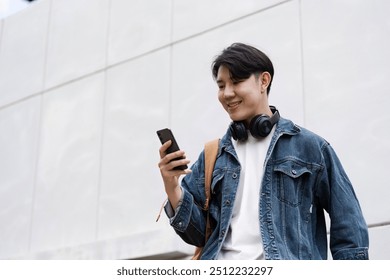 Young Student Smiling While Using Smartphone Outdoors, Wearing Headphones and Backpack, Casual Denim Jacket
