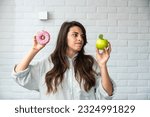 Young student woman doubts what to choose healthy food or sweets junk unhealthy food holding green apple and donut in hands standing at home in her living room. Hard choice concept