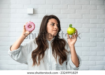 Young student woman doubts what to choose healthy food or sweets junk unhealthy food holding green apple and donut in hands standing at home in her living room. Hard choice concept