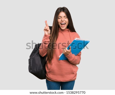 Young student woman holding notebooks with fingers crossing over isolated grey background