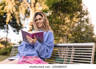 Young student woman holding personal diary sitting on bench in summer park, look happy and smiling. Blonde curly girl wear purple hoody, skirt, hold pink notebook, pink bag on bench. 