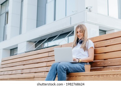 Young Student Woman Working At Laptop Sit Down On Bench Outside On A Urban City Street. Happy Lady Girl Drinking Coffee And Distance Learning, Education And Online Shopping.